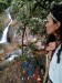 A woman looks away towards a rocky waterfall flowing into a small stream. On the rocky riverbank beside her are small offerings as part of a gratitude ritual to the water. thumbnail